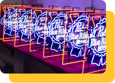 Glowing blue and orange Wholesale LED Neon Sign orders of Pabst Blue Ribbon Beer Signs stacked on assembly line.