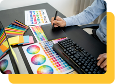 A graphic designer sits at their laptop with a rainbow of color charts across the desk to design LED Neon signs.